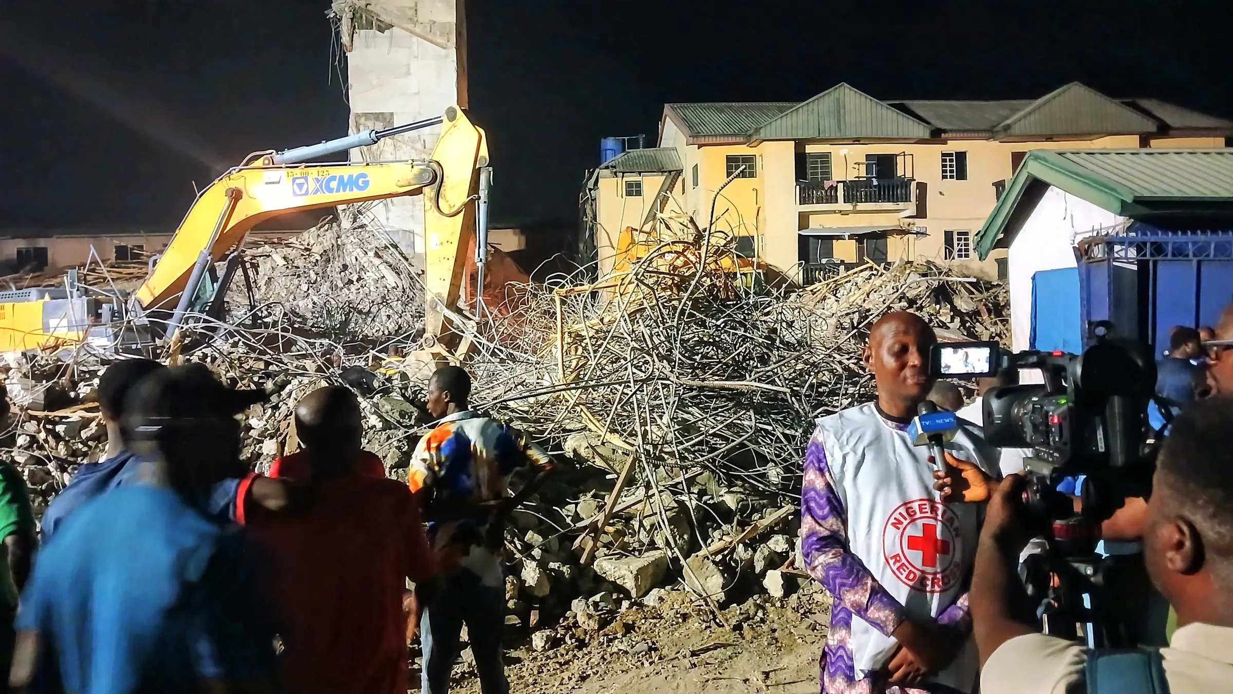 Rubble and twisted reinforcement bars of a collapsed building at night, with an excavator clearing debris