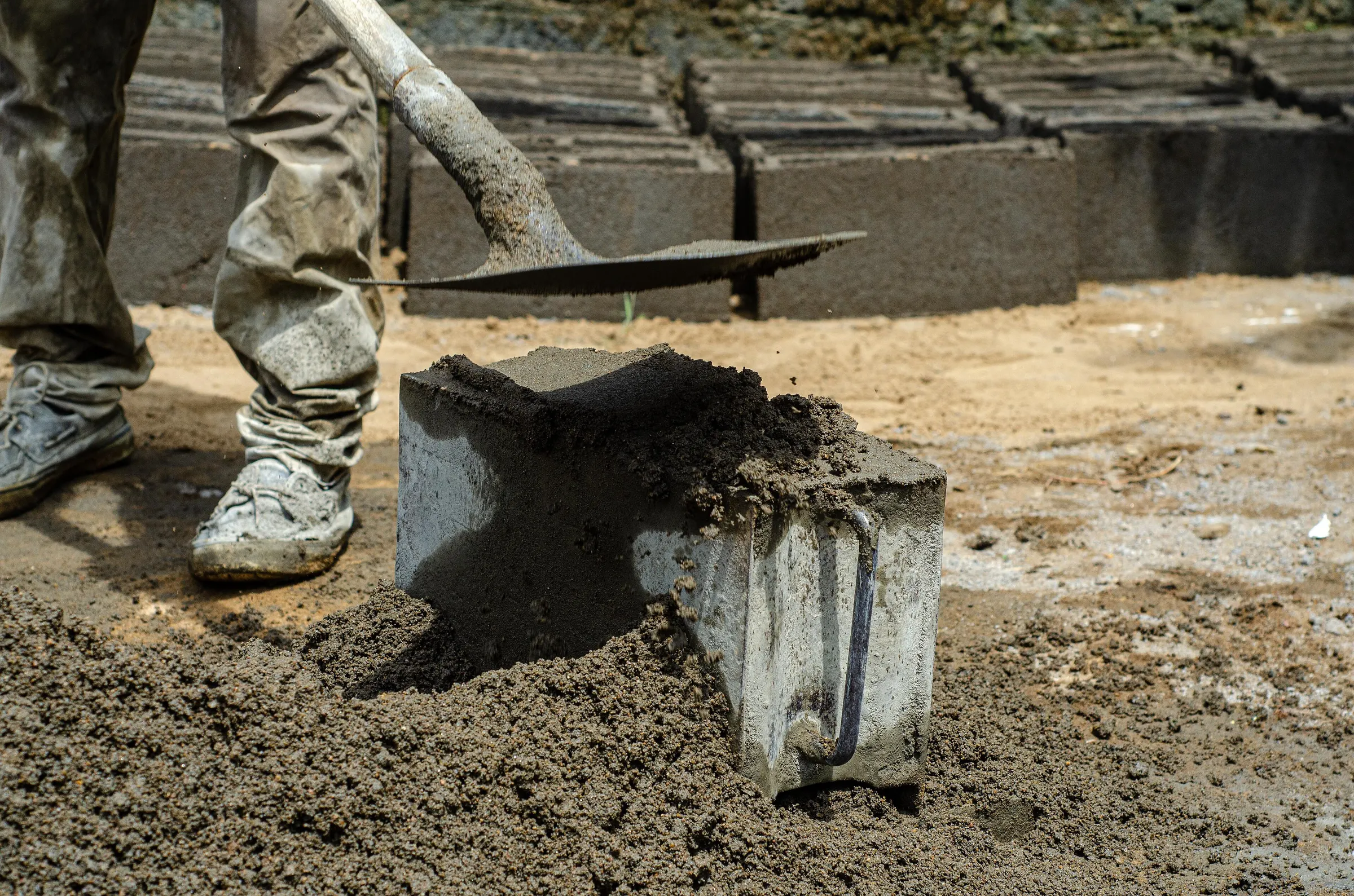 Shovel pressing cement mix into a block mould, with finished blocks behind
