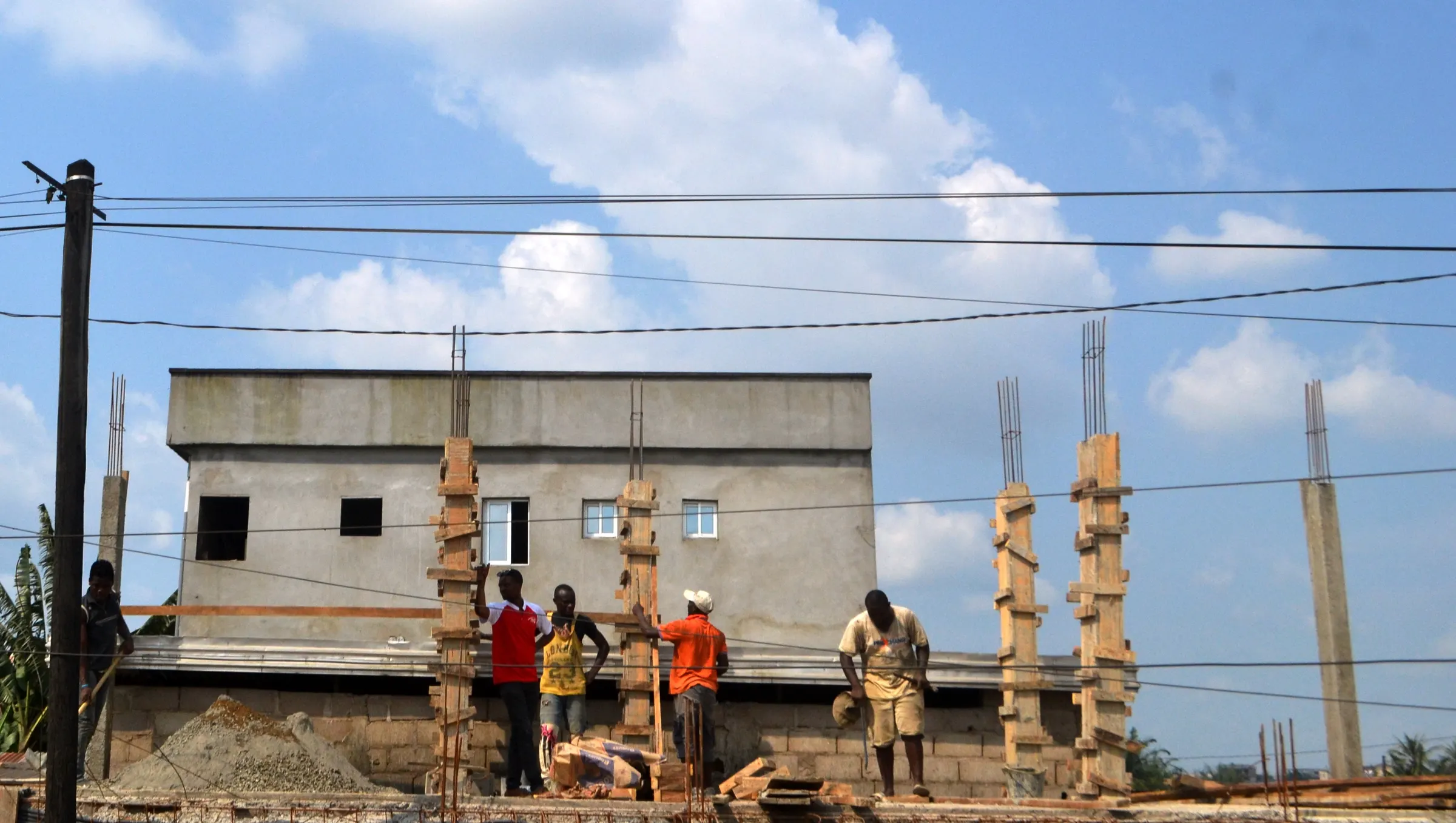 Workers on a building site with timber-shuttered columns and a two-storey block building behind