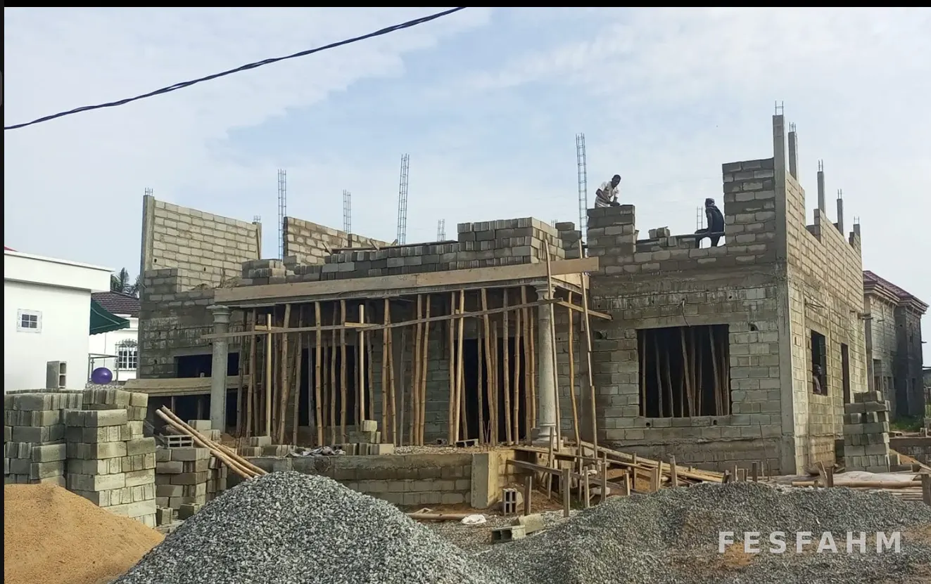 Cement-block house under construction with walls at beam level, propped formwork, starter bars and stacks of blocks, sand and gravel