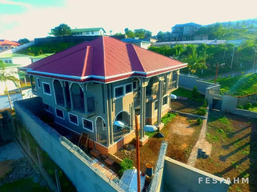 Aerial view of a completed two-storey villa with a red hipped roof and arched balconies inside a walled, landscaped compound